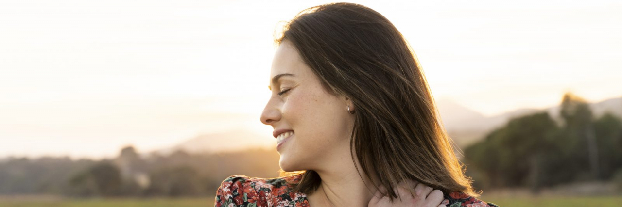 A white woman with brown hair wearing a floral blouse hugs herself outside.