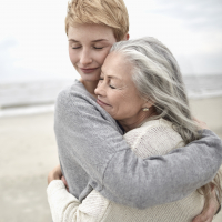 A person with short red hair hugs a woman with silver hair on the beach.