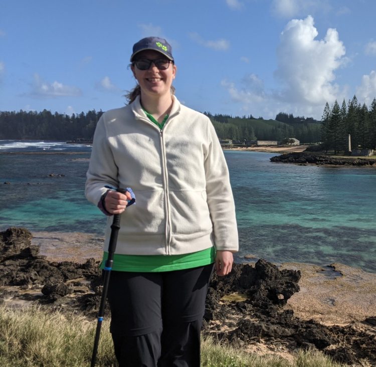 A woman wearing a white jacket, black pants, a navy blue baseball cap, and sunglasses who's holding a walking stick stands in front of a lake, smiling. 