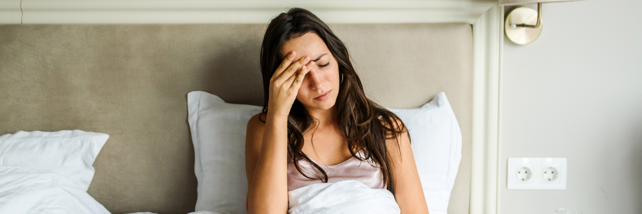 What Living With Long-Haul COVID-19 Really Feels Like A woman with brown hair sits in bed and covers up with a sheet while holding her head in pain.