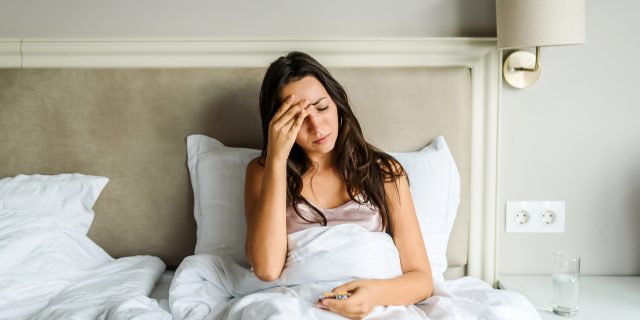 What Living With Long-Haul COVID-19 Really Feels Like A woman with brown hair sits in bed and covers up with a sheet while holding her head in pain.
