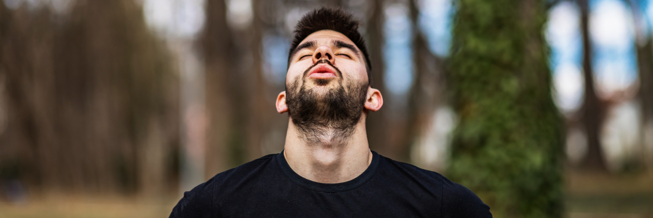 A man with dark brown hair wearing a black t-shirt tilts his head back and breathes while standing outside.