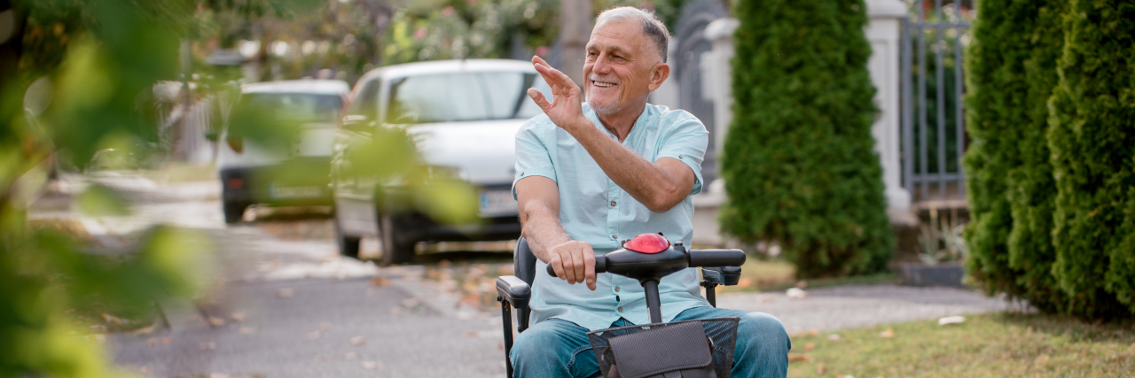 The High Cost of Mobility Equipment While Aging With Cerebral Palsy A man wearing a light blue shirt and blue jeans sits in a mobility scooter outside and waves.
