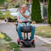 A man wearing a light blue shirt and blue jeans sits in a mobility scooter outside and waves.