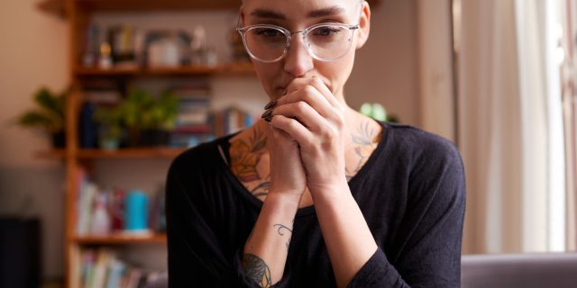 A person with short hair and glasses wearing a long-sleeved shirt holds their hands against their mouth while gazing ahead.