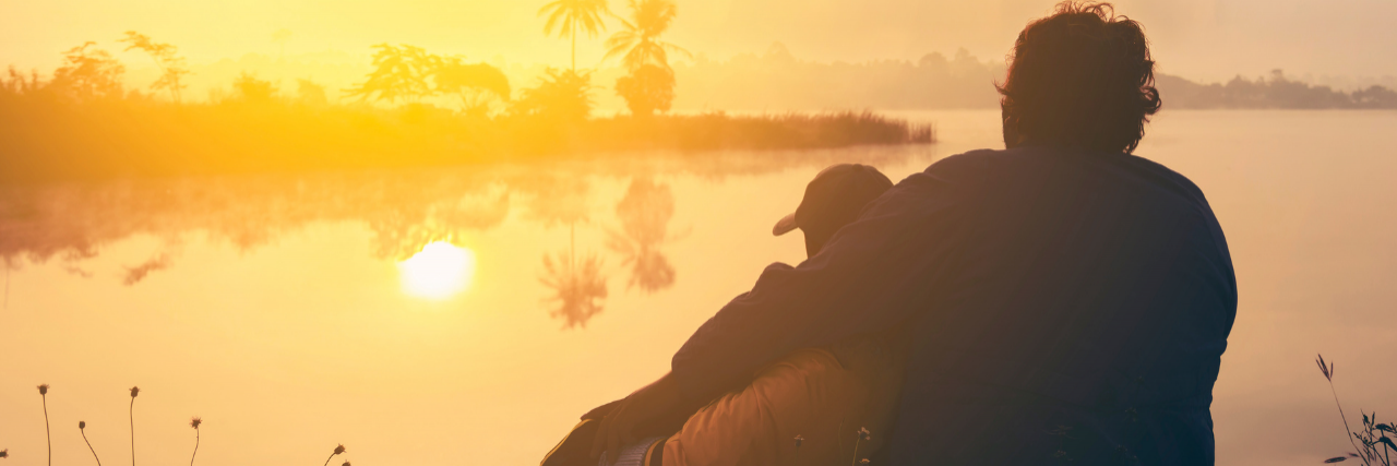 Two people sitting and embracing overlooking water and the sunset