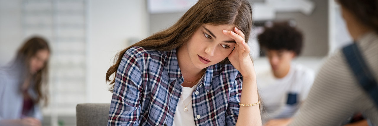 Being Told You're 'Too Young' to Have Fibromyalgia in Your 20s A young white woman with long brown hair wearing a long-sleeved blue plaid button-down and a white shirt writes in a classroom while cupping her head with her hand.