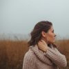 A brunette woman with brown eyes wearing a beige sweater stands in a field and looks to the side against a foggy sky.