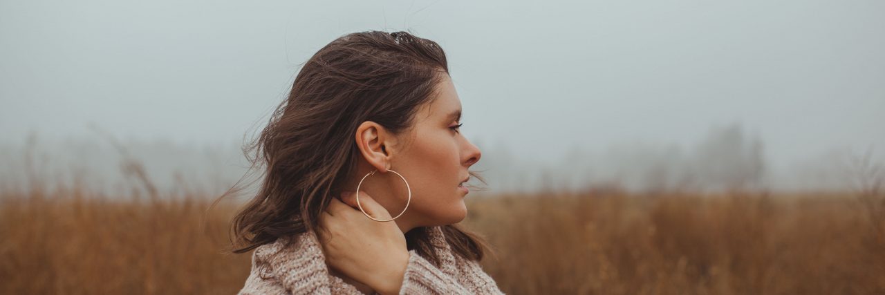 Learning to Embrace Self-Love After Years of Internalized Ableism A brunette woman with brown eyes wearing a beige sweater stands in a field and looks to the side against a foggy sky.