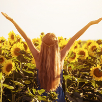 A blonde woman wearing a blue dress raises her arms in a field of sunflowers.