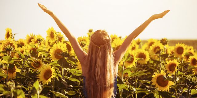 A blonde woman wearing a blue dress raises her arms in a field of sunflowers.