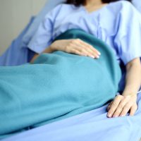 A woman wearing a blue hospital gown lies in a hospital bed with a blue blanket covering her.