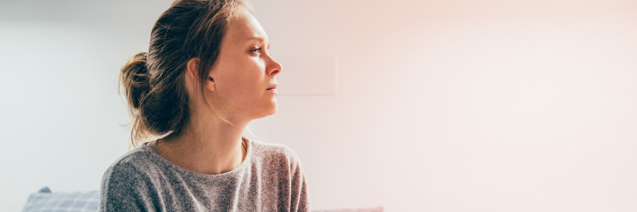 What I Want Myself to Know When My Chronic Pain Comes Back A woman with brown hair in a bun and a gray sweater sits on a bed and gazes into the distance. Her arms are crossed.