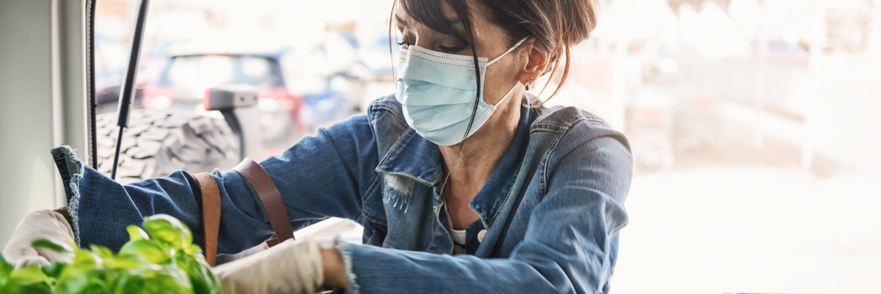 A woman with brown hair in a bun wearing a jean jacket and striped shirt loads a crate of groceries into her care while wearing a mask and gloves.