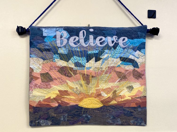 A handwoven tapestry embroidered with the word "Believe" along with a sunrise.