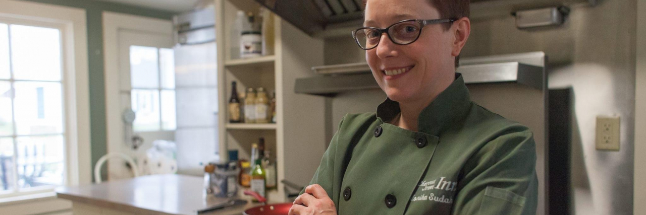 The Problematic Bullying In Cooking Shows Like 'Rat In The Kitchen' Photo of the contributor, Monika Sudakov, posing in a kitchen with a smile and her arms folded.
