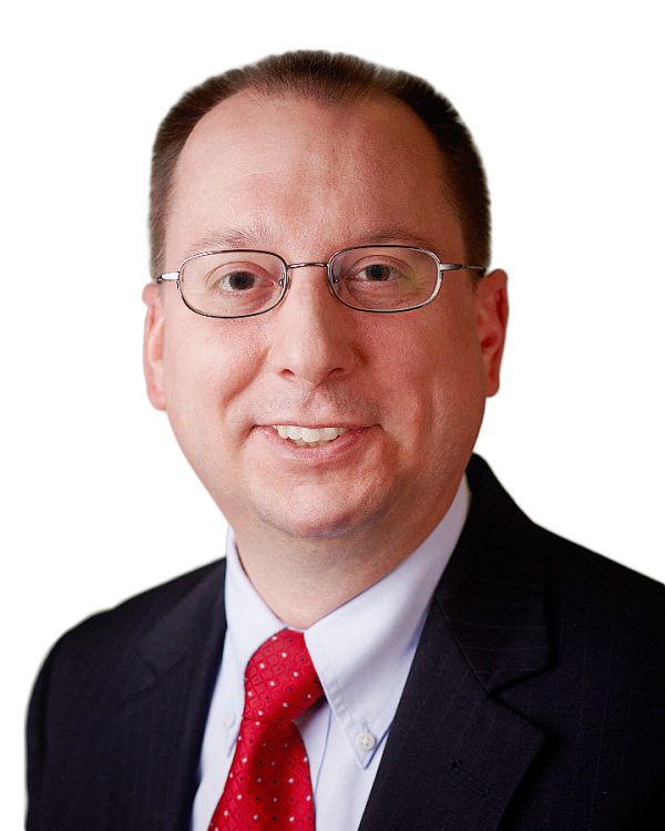 A white man with brown hair and brown eyes wearing glasses and a black suit with a red tie smiles. 