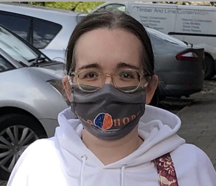 A white woman with brown hair wearing a gray face mask and a white hoodie stands in a parking lot. 