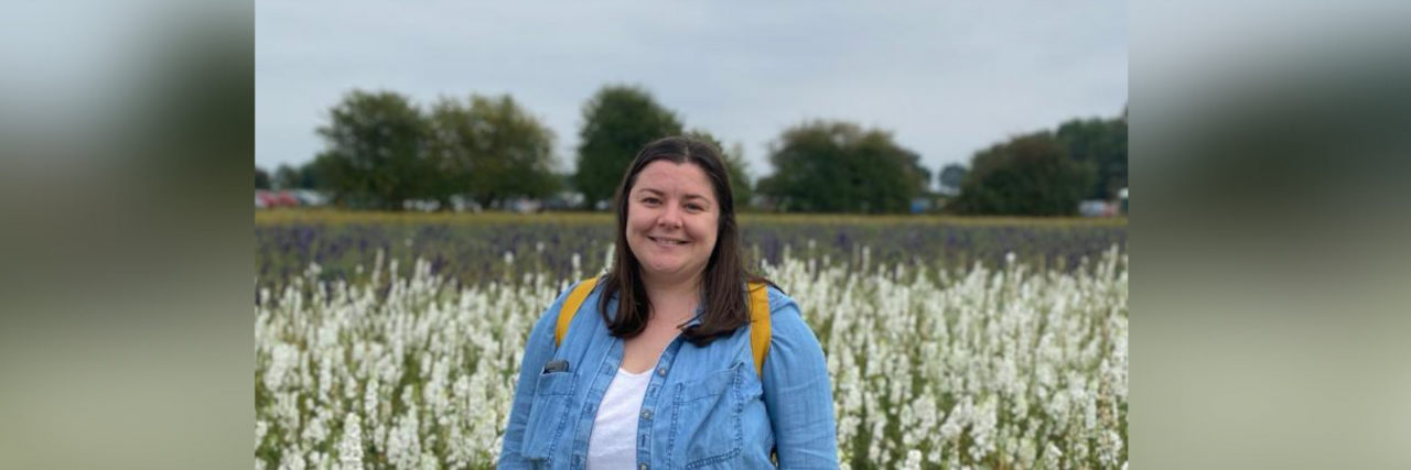 What Helps a Woman in Her Twenties With Depression A white woman with brown hair wearing a white shirt, coral patterned skirt, and jean jacket stands in a field of flowers.