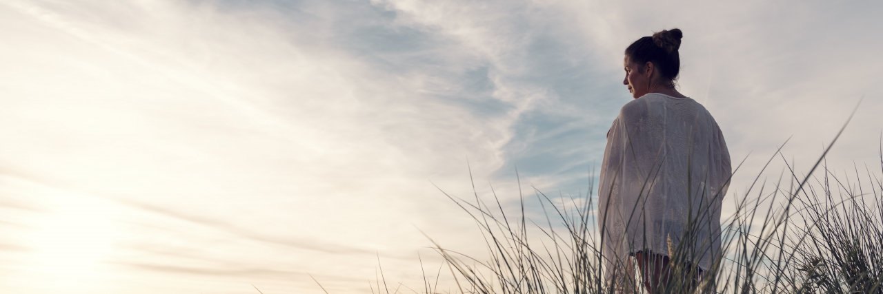 6 Ways Living With Migraine Can Affect Your Mental Health Woman standing on top of sand dunes at beach.