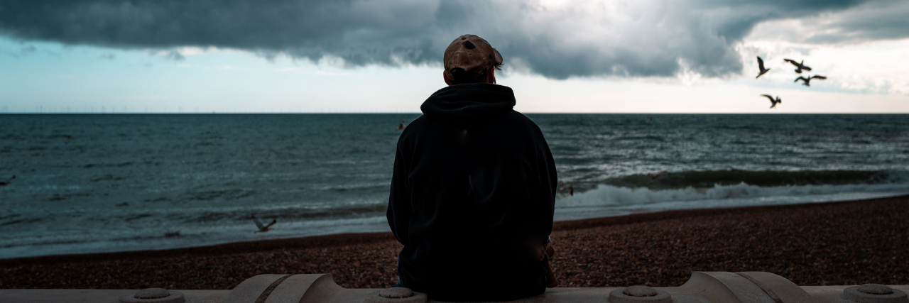 Back of a man sitting alone on the beach as a dark storm approaches
