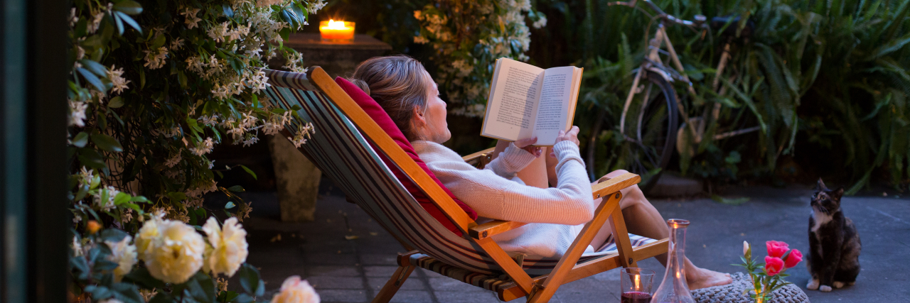 Self-Care Is Not Selfish: 10 Chronic Illness Self-Care Tips Woman relaxing on deck chair in back yard, reading a book with her cat watching.