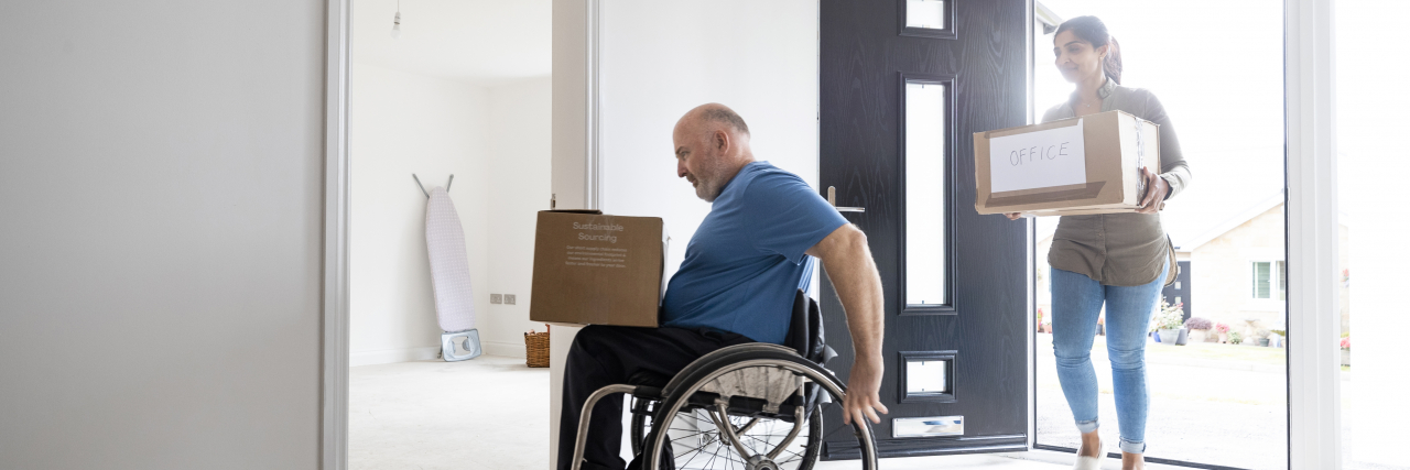 A mature couple carrying cardboard moving boxes. The man is a wheelchair user.