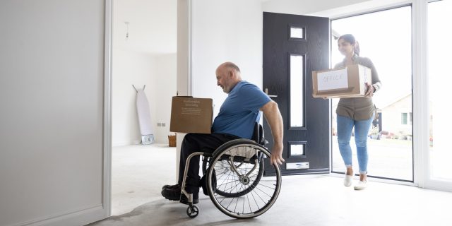 A mature couple carrying cardboard moving boxes. The man is a wheelchair user.
