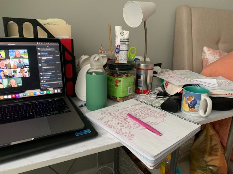 A desk with a notebook, waterbottle, protein snacks, laptop, mugs, and a lamp