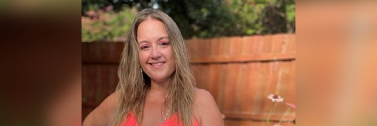 What Helps a Woman in Her Thirties With Complex PTSD A white woman with blonde hair wearing a coral dress poses in a garden with her hand on her hip.