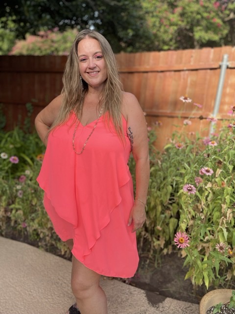 A white woman with blonde hair wearing a coral dress poses in a garden with her hand on her hip.