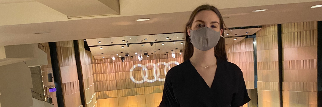 The Powerful Connection Between Disability and Music A photo of Carly Fox, a 20 year-old white woman, standing inside at the National Arts Centre of Canada in a deep blue wrap dress.