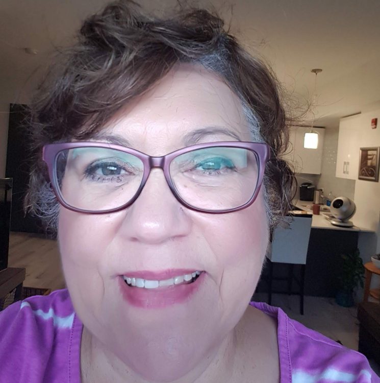 A white woman with short brown hair and glasses wearing a purple and white shirt smiles.