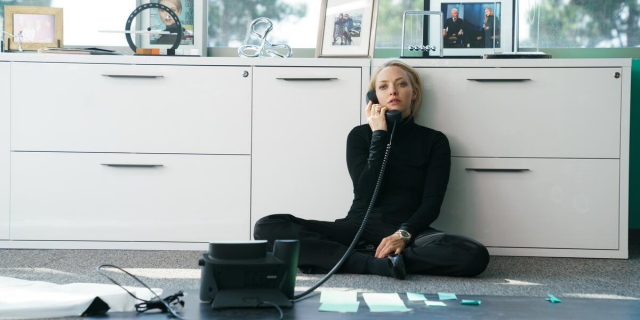 Amanda Seyfried in a black turtleneck sitting against a desk making a phone call