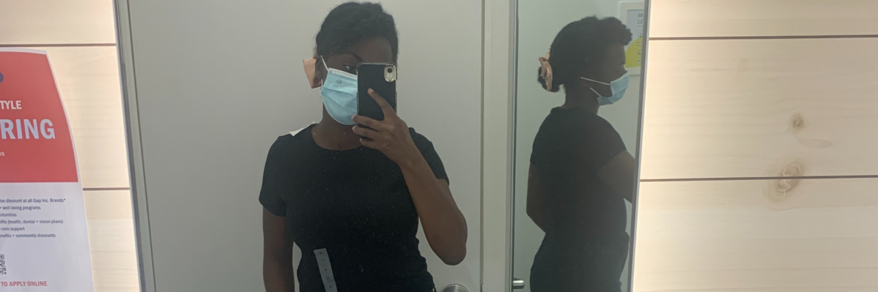 How to Buy New Clothes When You Struggle With Body Dysmorphia A girl looks in the mirror with her phone in a dressing room with a shirt and jeans on