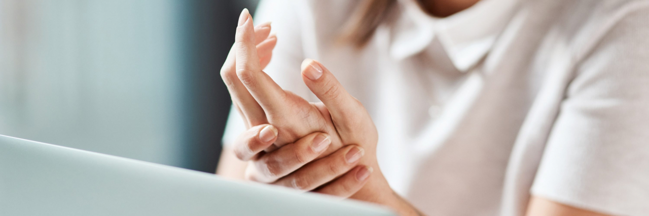 Losing Remission and Finding Gratitude With Arthritis A woman wearing a white short-sleeved blouse and sitting in front of a laptop holds her hand in pain.