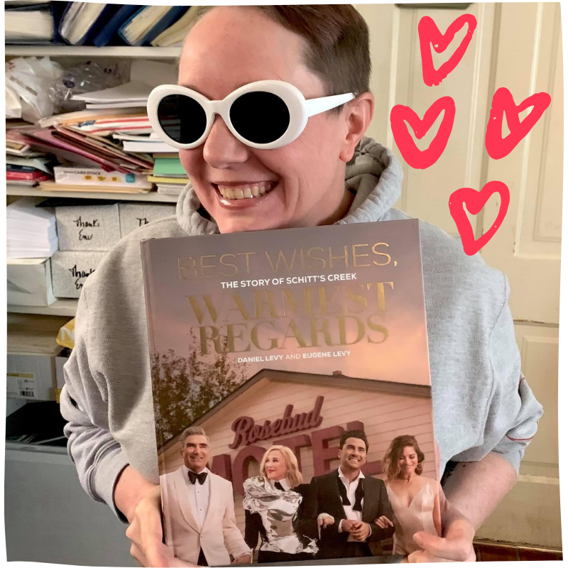 photo of Monika Sudakov holding a Schitt's Creek book, wearing white sunglasses and grinning