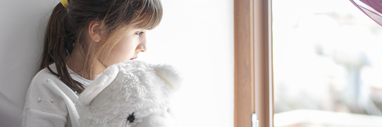 My Mother Lost Custody of Me Because She's Disabled A young girl with brown hair in a ponytail holds a white teddy bear while looking out a window.