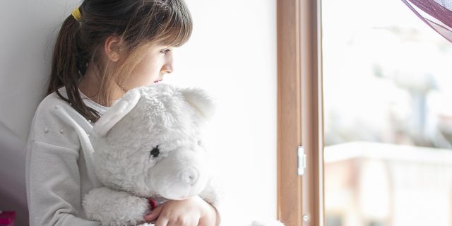 My Mother Lost Custody of Me Because of Her Disability A young girl with brown hair in a ponytail holds a white teddy bear while looking out a window.