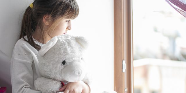 A young girl with brown hair in a ponytail holds a white teddy bear while looking out a window.