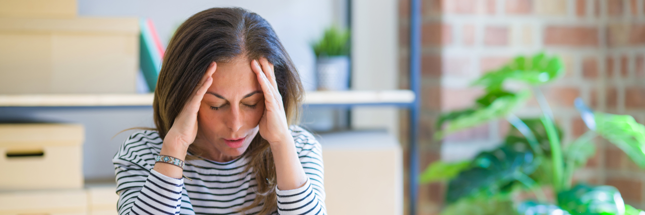 Long-Haul COVID-19 and Receiving a Behcet's Disease Diagnosis A woman swith brown hair wearing a black-and-white striped shirt holds her hands to her head during a headache.