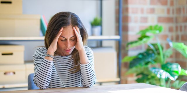 When I Learned My Long-Haul COVID-19 Led to Behcet's Disease A woman swith brown hair wearing a black-and-white striped shirt holds her hands to her head during a headache.