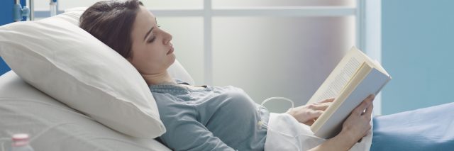 A woman with brown hair wearing a blue shirt lies in a hospital bed while reading a book.