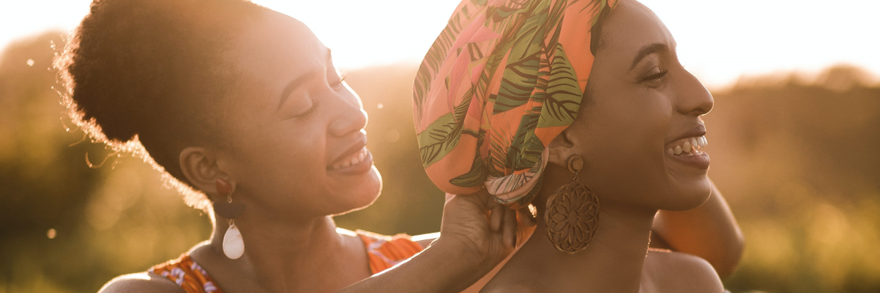 How Racism Impacts Black People With Alopecia photo of two women in low sunlight, one styling the other's hair under a headscarf