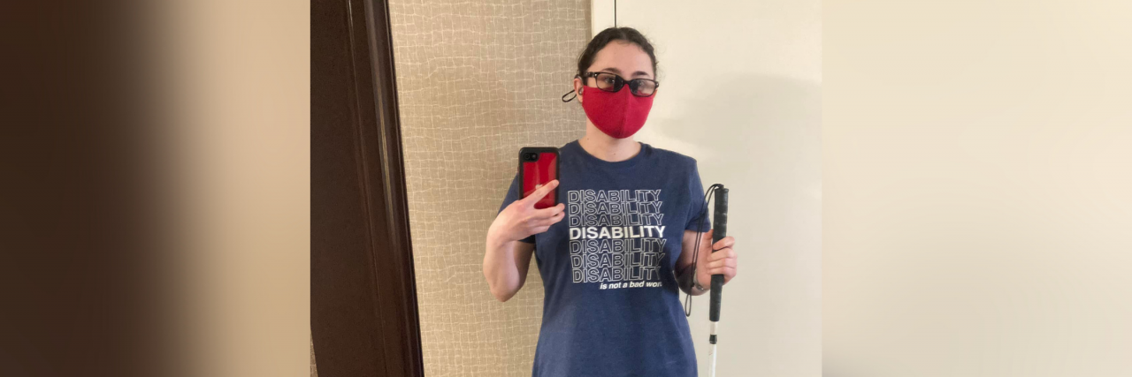 Disability Is Not a Bad Word Korrine standing up, taking a selfie in a stand up mirror. Her dark brown curly hair is tied back in a bun. She's wearing glasses, hearing aids, and a red mask. She's holding her white cane in one hand and her phone in the other. She's wearing a navy blue shirt that says DISABILITY is not a bad word, black leggings, and dark gray converse shoes.