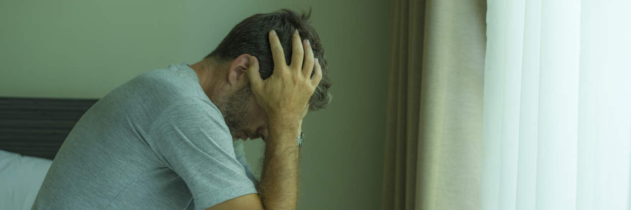 What I Want You to Know About How Autistic Shutdowns Affect Me A man with brown hair wearing a gray shirt and plaid pajama pants holds his head in his hands and sits on the edge of a bed.