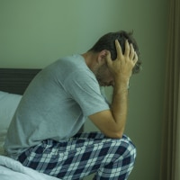 A man with brown hair wearing a gray shirt and plaid pajama pants holds his head in his hands and sits on the edge of a bed.
