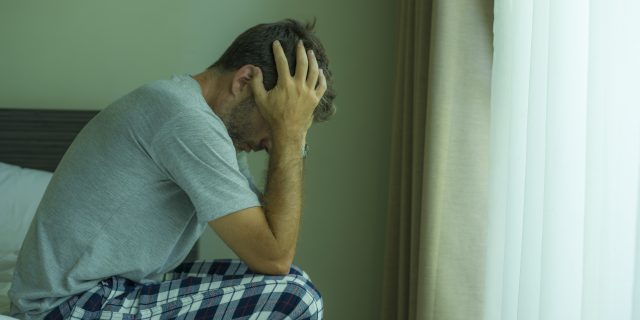 A man with brown hair wearing a gray shirt and plaid pajama pants holds his head in his hands and sits on the edge of a bed.