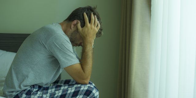 What I Want You to Know About How Autistic Shutdowns Affect Me A man with brown hair wearing a gray shirt and plaid pajama pants holds his head in his hands and sits on the edge of a bed.