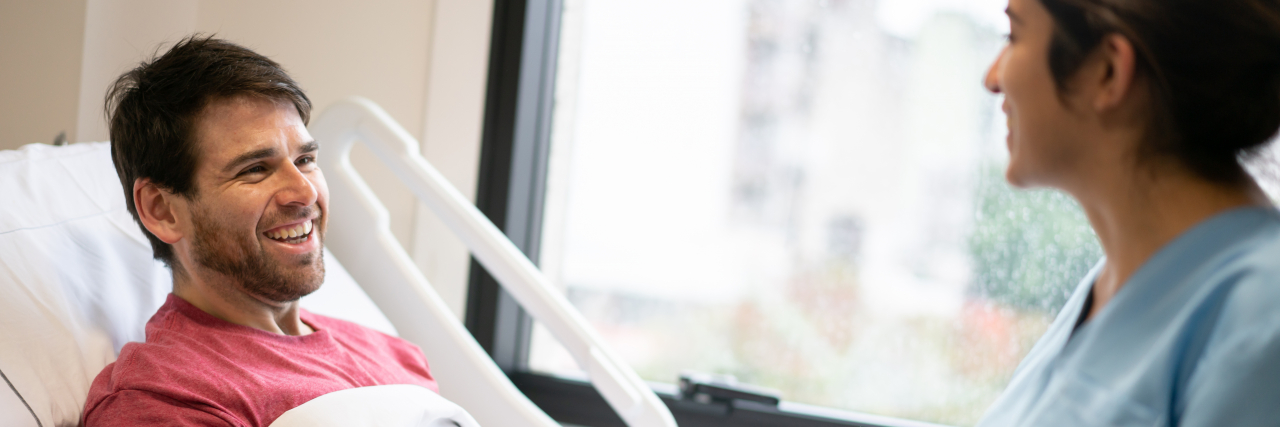 Finding Humor in Recovering From COVID-19 in the Hospital A smiling man with brown hair and a red shirt lies in a hospital bed while talking with a woman nurse.